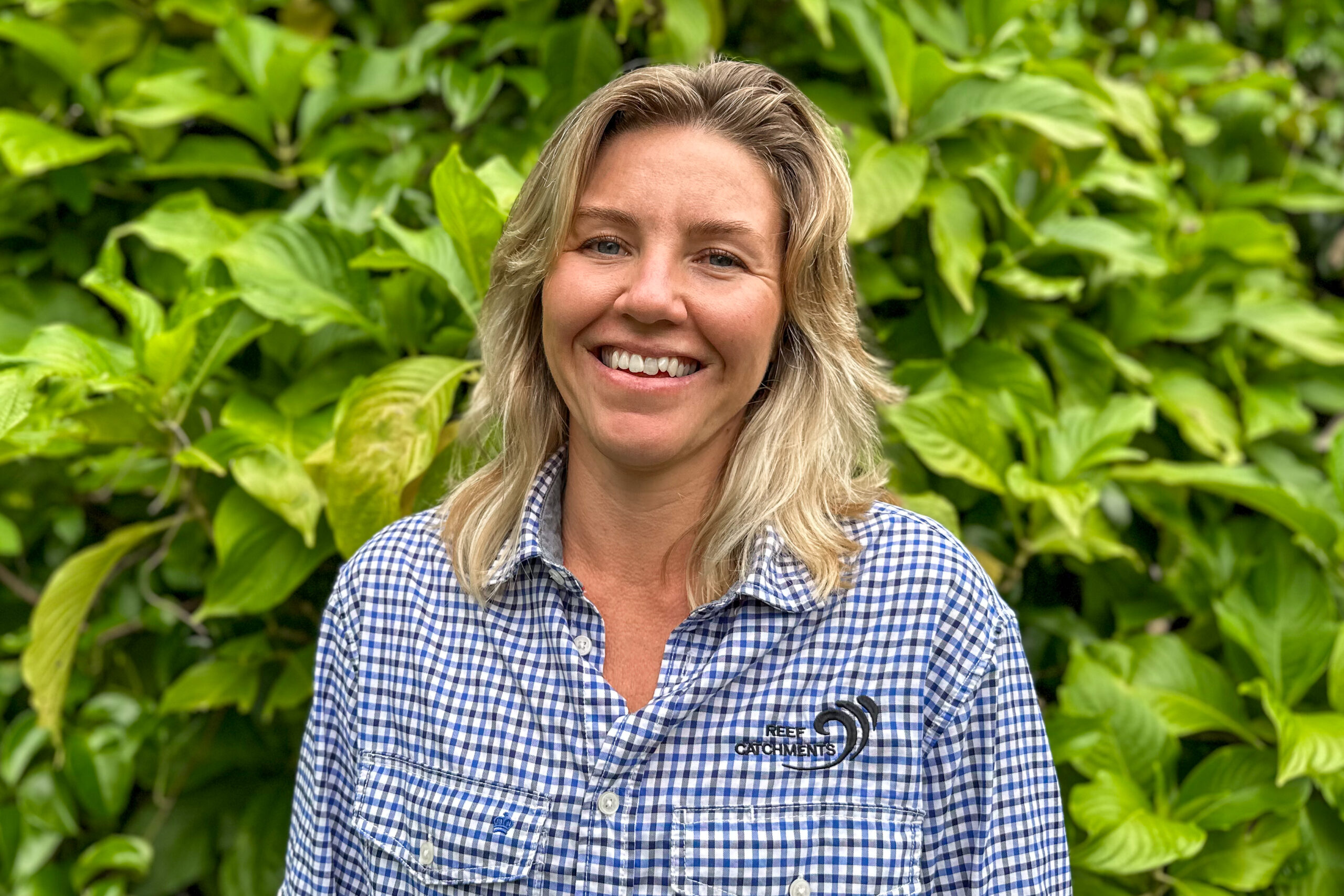 Holly Hayhurst headshot in front of a green leafy tree. Holly wears a blue and white checkered shirt with black Reef Catchments logo.